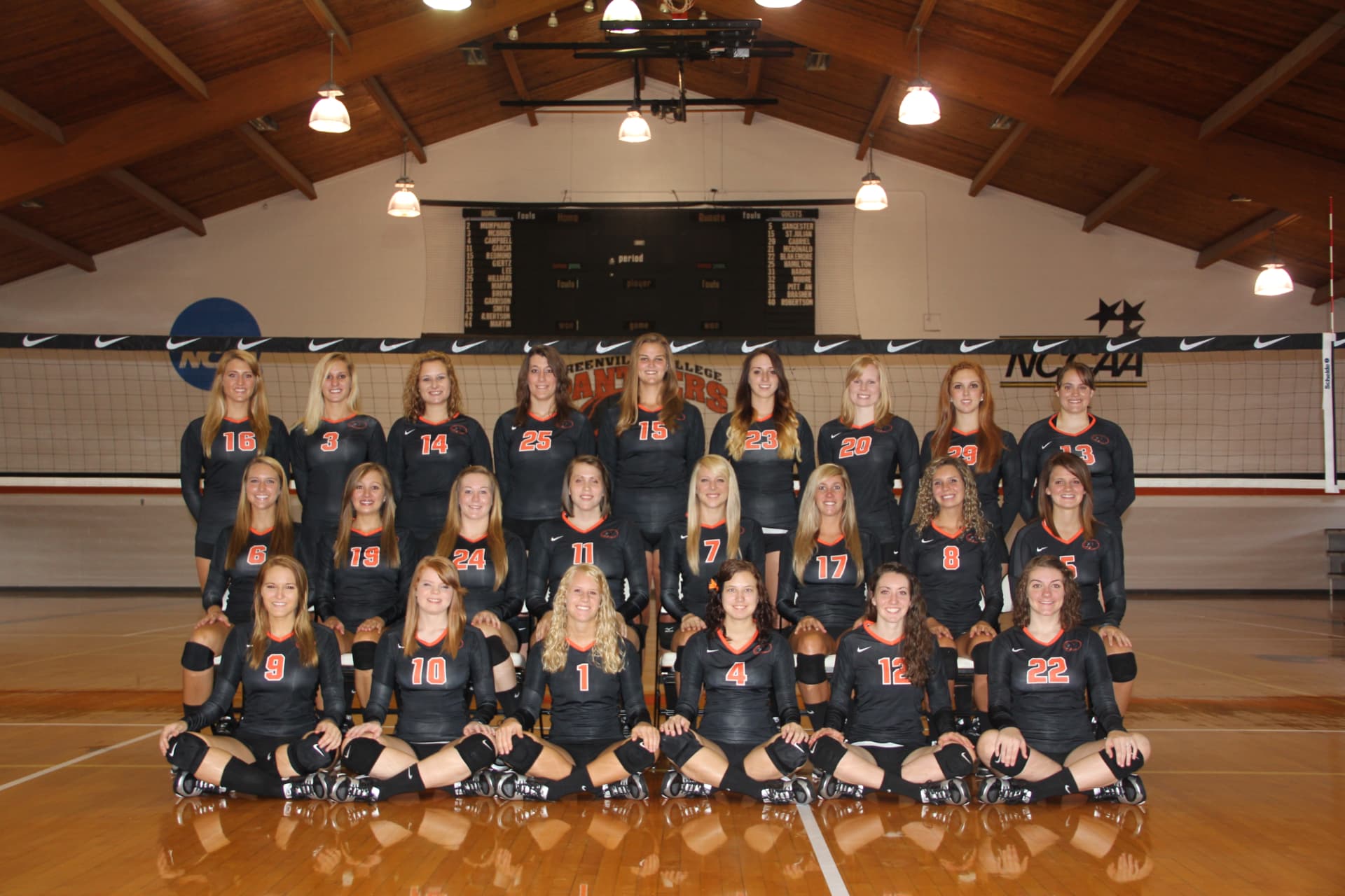 Greenville College women's volleyball team photo in black long-sleeve jerseys under the gym rafters.