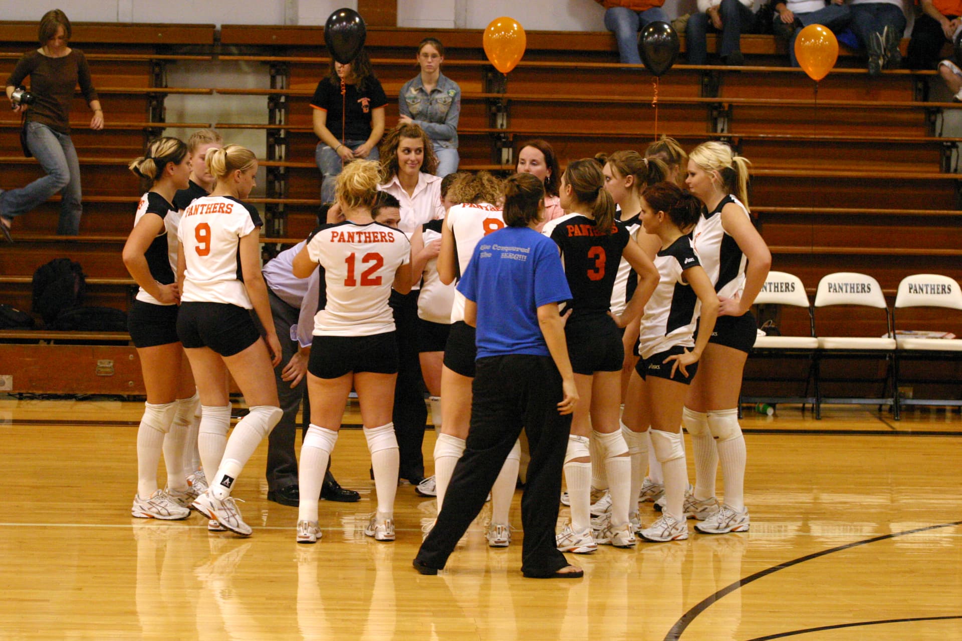 Greenville Panthers volleyball team huddling on the court with orange and black balloons in the stands.