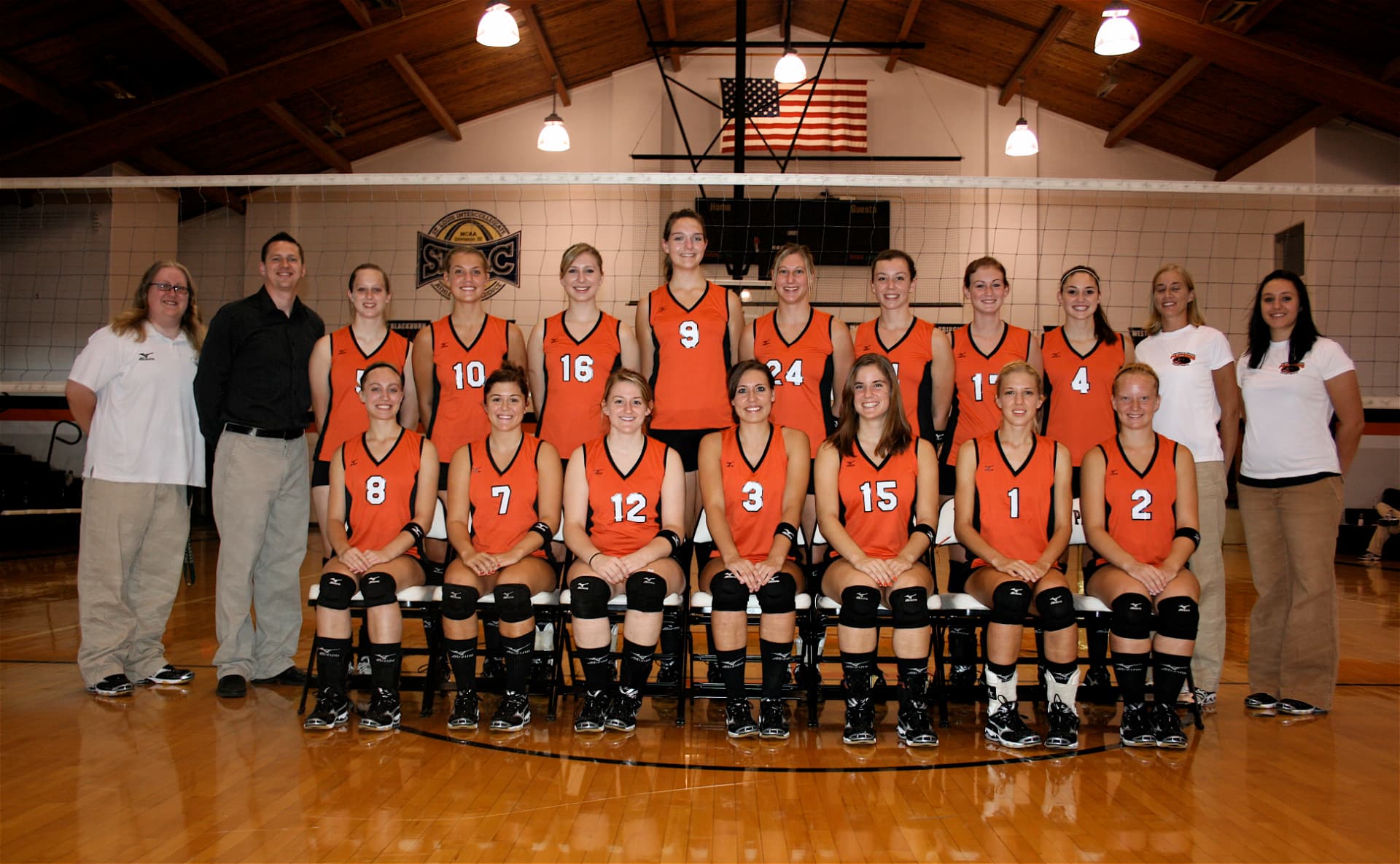 Greenville women's volleyball team photo in orange jerseys with coaches, in front of the net.